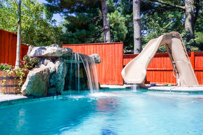 Mosquito B - DE - This professional installation features a multi-tier natural stone waterfall cascading into a gunite pool, with integrated artificial rock formations creating a grotto effect. The waterfall includes a spillway and multiple tiers, while a smooth concrete slide curves from the left side, descending into the pool. The project demonstrates exceptional craftsmanship with seamless integration of natural materials and modern water features in a private backyard setting surrounded by mature trees and a wooden fence.