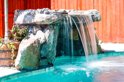 Mosquito C - DE - This custom outdoor living project features a multi-tier natural stone waterfall cascading into a gunite pool. The waterfall is constructed from large, textured boulders arranged in a tiered design, creating a serene cascade that flows into the clear blue pool water below. The surrounding area includes a wooden planter with vibrant flowers, enhancing the natural aesthetic of the backyard setting.