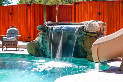 Mosquito E - DE - This custom installation features a multi-tier natural stone waterfall with smooth, cascading water flowing over textured boulders into a gunite pool. The waterfall includes a central spillway flanked by rock formations, creating a serene grotto effect. The surrounding area showcases quality craftsmanship with a wooden fence backdrop and comfortable poolside seating, enhancing the outdoor living space.