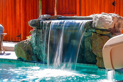 Mosquito F - DE - This custom installation features a multi-tier natural stone waterfall cascading into a gunite pool. The waterfall is constructed with large, irregularly shaped boulders that create a rugged, organic look, while the water flows smoothly over the stone surfaces. The pool itself has a clean, modern gunite finish with a gentle slope leading into the waterfall, creating a serene and inviting outdoor living space.