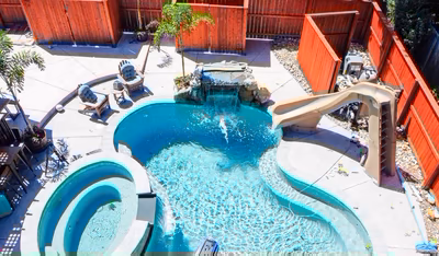 Mosquito J - DE - This custom installation features a natural stone waterfall cascading into a gunite pool with a built-in slide. The multi-tiered design includes a small grotto area and a separate spa section, all surrounded by a clean concrete deck. The project showcases high-quality craftsmanship with seamless integration of water features and landscaping in a private backyard setting.