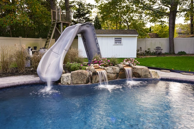 Nancy A - Lanoka Harbor, NJ - This professional installation features a large inflatable slide curving gracefully over a swimming pool, with two waterfall cascades integrated into natural stone boulders at the pool's edge. The slide's smooth, gray surface contrasts beautifully with the deep blue gunite pool below, while the surrounding landscape includes well-maintained grass, flowering shrubs, and a wooden play structure in the background. The craftsmanship demonstrates seamless integration of recreational and water features in a private backyard setting.