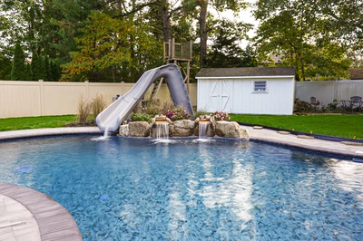 Nancy B - Lanoka Harbor, NJ - This professional installation features a sleek gray slide cascading directly into a gunite pool, flanked by natural stone waterfalls creating gentle cascades over river rocks. The multi-tiered water features demonstrate exceptional craftsmanship with precise stone placement and seamless integration of the slide structure. The project is set in a well-maintained backyard with a clean concrete deck surrounding the pool, landscaped with seasonal plants and backed by a white storage shed and wooden deck.