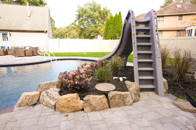 Nancy D - Lanoka Harbor, NJ - This professional installation features a large, gray plastic slide that curves gracefully into a gunite pool, creating a unique water feature. The pool is bordered by irregularly shaped natural stone boulders and lush landscaping with flowering plants, enhancing the natural aesthetic. The surrounding patio is constructed with light-colored pavers, and the background includes a well-maintained lawn, white fence, and comfortable outdoor seating area, showcasing high-quality craftsmanship and thoughtful design integration.
