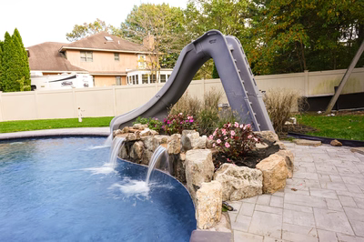 Nancy E - Lanoka Harbor, NJ - This professional installation features a dual gray slide waterfall system feeding into a gunite pool, with natural stone boulders creating a tiered waterfall effect. The project showcases high-quality craftsmanship through seamless integration of artificial rock formations and smooth concrete surfaces, set in a well-maintained backyard with manicured grass and flowering plants.