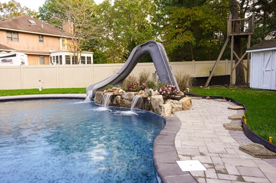 Nancy F - Lanoka Harbor, NJ - This professional installation features a sleek grey slide that transitions seamlessly into a gunite pool, surrounded by a natural stone waterfall with multiple tiers. The waterfall uses large river rocks to create a cascading water effect that flows into the pool, enhancing both aesthetics and functionality. The project is set in a well-maintained backyard with a clean concrete patio, lush green grass, and surrounding trees, creating an inviting outdoor living space.