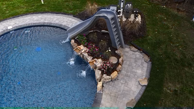 Nancy G - Lanoka Harbor, NJ - This professional installation features a gunite pool with a custom gray slide waterfall that cascades into the pool. The waterfall is constructed with durable synthetic rock and includes a built-in staircase for access. The pool deck is finished with interlocking brick pavers and accented with natural stone boulders and flowering plants, creating a cohesive and inviting backyard retreat in Lanoka Harbor, NJ.
