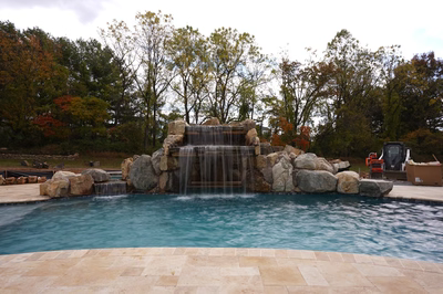 Ndakaini A - Owings Mill, MD - This custom outdoor living project features a multi-tier natural stone waterfall cascading into a gunite pool, surrounded by large river boulders that enhance the natural aesthetic. The waterfall includes a central spillway flanked by tiered stone ledges, creating a soothing water feature that flows seamlessly into the deep blue pool below. The surrounding area is landscaped with autumn-colored trees, providing a serene backdrop to this high-quality installation in Owings Mill, MD.
