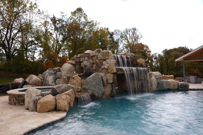 Ndakaini C - Owings Mill, MD - This custom outdoor living project features a multi-tier natural stone waterfall cascading into a gunite pool. The waterfall is constructed with large, irregularly shaped boulders arranged in a tiered design, creating a natural, grotto-like appearance. The pool itself has a smooth, gunite finish with a clean, inviting blue water surface, accented by additional large boulders along the pool's edge for a rustic, natural aesthetic. The setting is a well-maintained backyard with autumn-colored trees in the background, providing a serene and luxurious outdoor relaxation space.