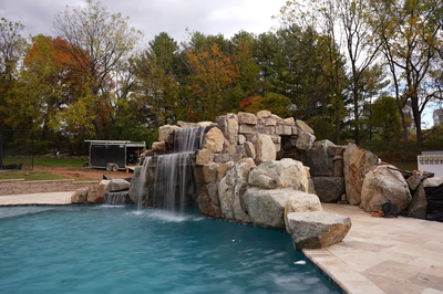 Ndakaini E - Owings Mill, MD - This custom outdoor living project features a multi-tier natural stone waterfall cascading into a gunite pool. The waterfall is constructed from large, irregularly shaped boulders arranged to create a naturalistic flow, with water gently cascading over each tier. The pool itself has a clean, freeform edge and is surrounded by a stone deck, enhancing the natural aesthetic of the setting. The surrounding landscape includes mature trees with autumn foliage, providing a serene and private backdrop for this luxurious backyard oasis.