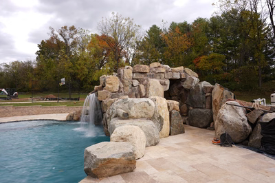Ndakaini F - Owings Mill, MD - This custom outdoor living project features a multi-tier natural stone waterfall cascading into a gunite pool. The waterfall is constructed from large, irregularly shaped boulders arranged to create a rugged, organic flow, with water gently spilling over each tier into the pool below. The surrounding area includes a spacious, light-colored travertine patio that complements the natural stone elements, creating a harmonious and relaxing backyard setting surrounded by autumn-colored trees.