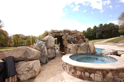 Ndakaini G - Owings Mill, MD - This custom outdoor living project features a natural stone grotto with a built-in pool and waterfall. The structure uses large, irregularly shaped boulders and natural stone to create an organic, rustic aesthetic. The pool has a smooth, gunite finish with a gently sloping edge, and the waterfall cascades down from a stone ledge into the pool below. The setting is surrounded by a well-maintained garden with trees and a paved walkway leading to additional outdoor spaces.