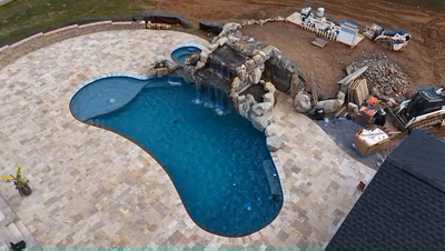 Ndakaini I - Owings Mill, MD - This professionally installed outdoor living project features a custom natural stone waterfall cascading into a gunite pool with a smooth, freeform edge. The waterfall is constructed with large, irregularly shaped boulders creating a natural, tiered cascade that flows into the deep blue pool below. The surrounding deck is finished with large, light-colored flagstone tiles that complement the earthy tones of the stone waterfall and create a luxurious, spa-like atmosphere in the Owings Mill backyard setting.