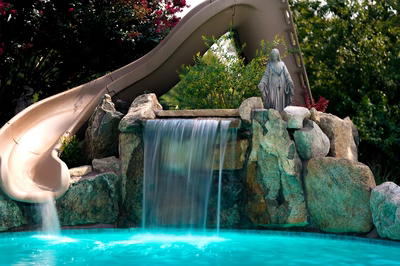 Rainbow B - Paulsboro, NJ - This professional installation features a multi-tier natural stone waterfall cascading into a gunite pool, integrated with a smooth concrete slide on one side. The waterfall uses large river rocks as accents and includes a serene statue positioned atop the rock formation. The craftsmanship showcases precise stonework and seamless integration of water features within a lush, landscaped backyard setting in Paulsboro, NJ.