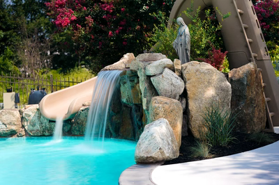 Rainbow C - Paulsboro, NJ - This professional installation features a multi-tier natural stone waterfall cascading into a gunite pool, with large river boulders creating a rugged, organic look. The waterfall includes an integrated concrete slide that curves down into the pool, enhancing both aesthetics and functionality. Surrounded by lush landscaping with flowering shrubs and a serene statue, the project demonstrates exceptional craftsmanship and attention to detail in creating a tranquil backyard oasis.