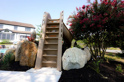 Rainbow G - Paulsboro, NJ - This professional installation features a custom concrete slide with perforated sides for drainage, integrated into a natural stone landscape. The slide is flanked by large, irregularly shaped boulders that blend seamlessly with the surrounding garden. The project demonstrates high-quality craftsmanship with clean concrete finishes and thoughtful integration of natural elements, creating an inviting backyard retreat adjacent to a swimming pool.