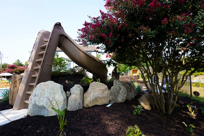 Rainbow H - Paulsboro, NJ - This custom rock slide features a smooth, curved plastic slide integrated with natural stone boulders at its base. The slide structure is supported by a sturdy concrete frame with multiple steps leading up to the top. The surrounding area is landscaped with mulch, small plants, and a flowering tree with pink blossoms, creating a serene backyard setting. The craftsmanship combines modern play equipment with natural elements for an inviting outdoor space.