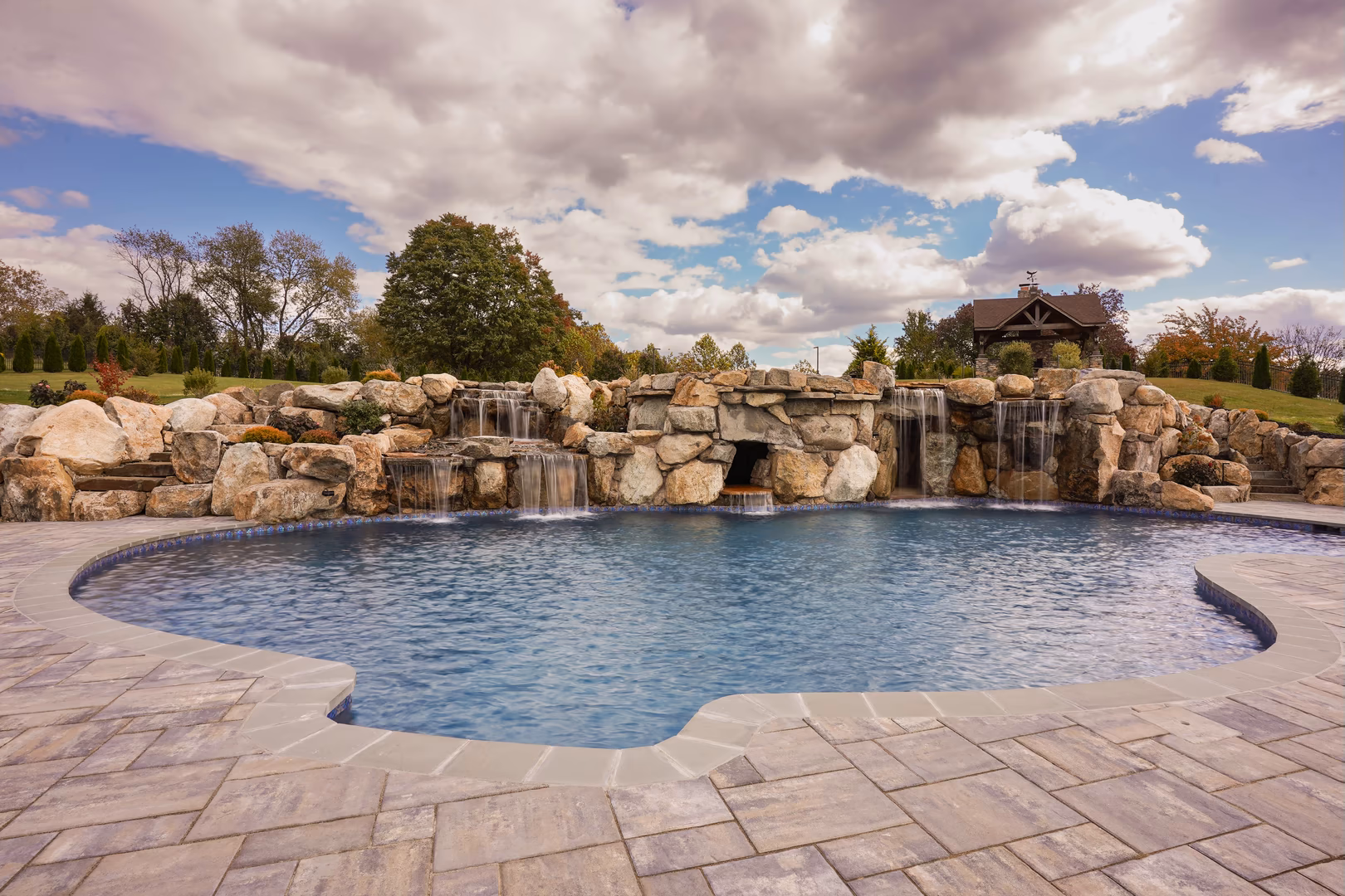 Sandor A - Finksburg, MD - This custom installation features a multi-tier natural stone waterfall cascading into a gunite pool with a smooth, inviting surface. The waterfall is constructed from large, irregularly shaped boulders arranged to create a natural, flowing cascade, with multiple tiers and spillways feeding into the central pool. The surrounding area is paved with large, light-colored stone tiles, enhancing the upscale, serene atmosphere of the backyard setting.