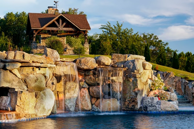 Sefinen D - Finksburg, MD - This professionally constructed outdoor living project features a multi-tier natural stone waterfall cascading into a gunite pool. The waterfall is built with large, irregularly shaped boulders arranged in a tiered design, creating a dynamic water feature with multiple spillways. The surrounding landscaping includes manicured grass, mature trees, and a wooden gazebo structure, creating a serene and luxurious backyard environment.