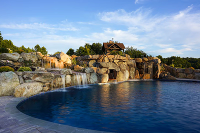 Sefinen C - Finksburg, MD - This custom installation features a multi-tier natural stone waterfall cascading into a gunite pool, with large boulders integrated into the design for a rustic, natural look. The waterfall includes multiple levels of cascading water, creating a soothing, tranquil atmosphere. The surrounding area is landscaped with additional boulders and greenery, enhancing the serene outdoor living space in a private backyard setting.
