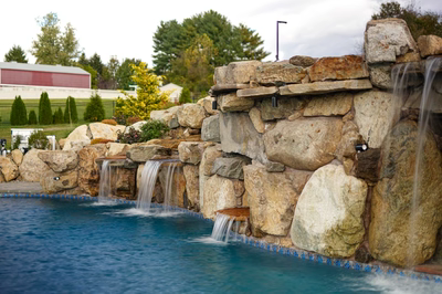 Sefinen F - Finksburg, MD - This custom installation features a multi-tier natural stone waterfall cascading into a gunite pool. The waterfall is constructed from large, irregularly shaped boulders arranged in a tiered design, creating multiple small cascades that flow into the pool below. The pool itself has a clean gunite finish with a blue tile accent along the edge, complementing the natural stone elements. The surrounding area includes additional boulders and landscaped greenery, enhancing the natural, rustic aesthetic of the outdoor living space in Finksburg, MD.