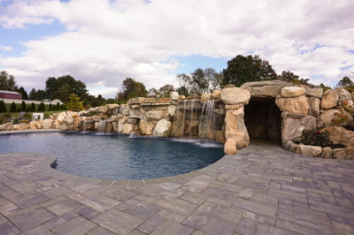 Sefinen H - Finksburg, MD - This custom installation features a multi-tier natural stone waterfall cascading into a gunite pool, with large boulders integrated into the design creating a grotto-like feel. The waterfall includes multiple spillways and cascades that flow into the pool, surrounded by meticulously placed stone pavers. The setting is a well-maintained backyard with mature trees and a serene atmosphere, showcasing high-quality craftsmanship and attention to detail in both the water features and surrounding landscaping.