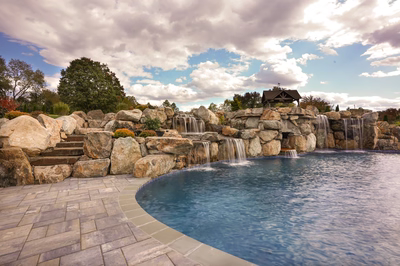 Sefinen B - Finksburg, MD - This custom installation features a multi-tier natural stone waterfall cascading into a gunite pool, with large boulders integrated into the design for a rustic, natural look. The waterfall includes multiple spillways and cascades, creating a soothing water feature that flows into the deep, inviting pool. The surrounding area is landscaped with additional boulders and a stone patio, enhancing the serene backyard setting under a partly cloudy sky.