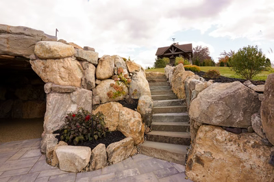 Sefinen I - Finksburg, MD - This custom outdoor living project features a natural stone grotto with a tiered waterfall cascading down rugged boulders. The construction showcases high-quality craftsmanship using large, irregularly shaped stones that create a rustic, organic feel. The setting includes a paved walkway leading up to a wooden house with a prominent weathervane, surrounded by well-maintained landscaping with shrubs and grass under a partly cloudy sky.