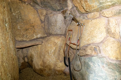 Sefinen N - Finksburg, MD - This custom shower installation features rough-hewn natural stone walls creating an authentic grotto atmosphere. Exposed copper piping and fittings provide both functionality and rustic charm, with a handheld showerhead mounted on a flexible hose for versatile use. The construction showcases excellent craftsmanship with precise stonework and durable metal components integrated seamlessly into the natural setting.