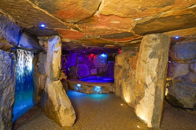 Sefinen P - Finksburg, MD - This custom installation features a multi-tier natural stone grotto with a prominent cascading waterfall on the left, flowing into a gunite pool with integrated LED lighting that creates a striking blue glow. The space includes a separate stone-lined spa area with additional lighting, surrounded by large, strategically placed boulders that enhance the natural, cave-like ambiance. The craftsmanship showcases seamless integration of natural materials and modern lighting to create an inviting, luxurious retreat.