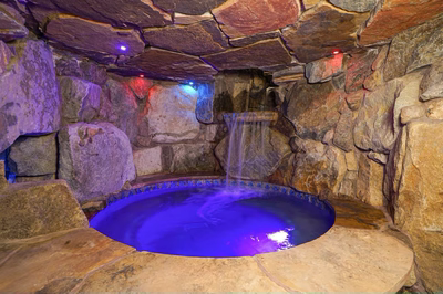 Sefinen S - Finksburg, MD - This custom installation features a natural stone waterfall cascading from a tiered rock formation into a gunite pool with a striking blue LED illumination. The surrounding area is accented with large, irregularly shaped boulders that enhance the natural, grotto-like ambiance. The craftsmanship showcases seamless integration of stone materials and precise water flow design, creating a relaxing and visually appealing outdoor living space.