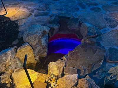 Sefinen T - Finksburg, MD - This custom installation features a natural stone waterfall with a smooth, curved spillway illuminated by vibrant blue LED lighting. The structure uses large, irregularly shaped boulders and flagstone accents, creating a rustic yet modern aesthetic. The craftsmanship demonstrates precise stonework and thoughtful integration of lighting to enhance the nighttime ambiance in this Finksburg, MD outdoor living space.