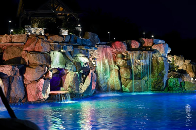 Sefinen U - Finksburg, MD - This professionally installed multi-tier waterfall features vibrantly colored natural stone boulders arranged in a cascading design, with illuminated water flowing over each tier into a deep blue gunite pool below. The installation demonstrates exceptional craftsmanship through precise stone placement and integrated lighting that enhances both the rock textures and water movement, creating a dramatic nighttime focal point in a landscaped outdoor setting.