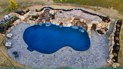 Sefinen V - Finksburg, MD - This professionally installed outdoor living project features a custom natural stone waterfall cascading into a gunite pool. The multi-tiered waterfall is constructed from large, irregularly shaped boulders that create a naturalistic, freeform design, with water flowing over multiple levels before reaching the pool. The surrounding area is paved with interlocking stone tiles, and the pool deck is bordered by additional boulders and low-maintenance landscaping, creating a serene and inviting backyard retreat in Finksburg, MD.