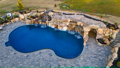 Sefinen W - Finksburg, MD - This custom installation features a multi-tier natural stone waterfall cascading into a gunite pool with a smooth, freeform edge. The waterfall includes multiple levels of cascading water, integrated with large, irregularly shaped boulders that create a natural, grotto-like appearance. The surrounding area is paved with interlocking slate tiles, enhancing the rustic aesthetic and providing a durable, low-maintenance surface.
