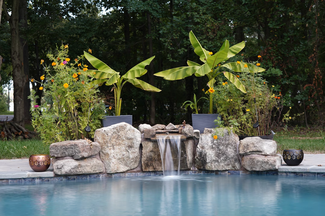 Swadini A - Grasonville, MD - This professionally installed natural stone waterfall features a multi-tiered cascade that flows gracefully into a gunite pool. The waterfall is constructed with large, irregularly shaped boulders, creating a rustic and organic look that blends seamlessly with the surrounding tropical plants, including banana trees and flowering shrubs in decorative pots. The setting is a tranquil backyard oasis in Grasonville, MD, showcasing high-quality craftsmanship and attention to detail in both the water feature and landscaping design.