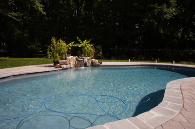 Swadini D - Grasonville, MD - This professional installation features a natural stone waterfall cascading gently into a gunite pool, accented by strategically placed boulders that enhance the natural aesthetic. The pool's clean lines and smooth surface reflect the surrounding greenery, while the surrounding patio area is constructed with durable stone pavers. The setting is a well-maintained backyard in Grasonville, MD, with lush landscaping and mature trees providing a serene backdrop.
