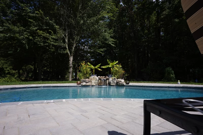 Swadini I - Grasonville, MD - This professional installation features a multi-tier natural stone waterfall cascading into a gunite pool, with large river boulders integrated into the design for a rustic aesthetic. The waterfall includes a tiered cascade with a small spillway, creating a soothing water feature that flows into the clear blue pool water. The surrounding area is landscaped with lush greenery and tropical plants, enhancing the serene backyard setting in Grasonville, MD.