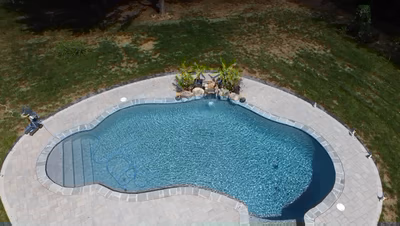 Swadini J - Grasonville, MD - This professional installation features a sleek gunite pool with a gracefully curved design, surrounded by a durable paver deck. A natural stone waterfall cascades gently from a rocky outcrop, integrating seamlessly with the pool's edge. The surrounding area is landscaped with well-maintained grass and tropical plants, creating a serene backyard retreat.