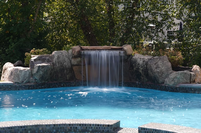 Yudaki A - Chalfont, PA - This custom installation features a multi-tier natural stone waterfall cascading into a gunite pool. The waterfall is constructed with large, irregularly shaped boulders that create a rugged, organic look, while the pool itself has a clean, smooth finish with mosaic tile accents along the edges. The setting is a well-maintained backyard with lush greenery providing a serene backdrop, enhancing the tranquil atmosphere of the outdoor living space.