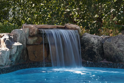 Yudaki B - Chalfont, PA - This custom installation features a multi-tier natural stone waterfall cascading into a gunite pool. The waterfall is constructed using large, irregularly shaped boulders arranged in a tiered design, creating a natural, freeform appearance. The pool itself has a clean, smooth gunite finish with a mosaic tile border, enhancing the luxurious and serene atmosphere of the outdoor living space.