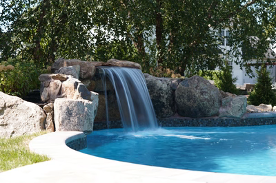 Yudaki C - Chalfont, PA - This custom installation features a natural stone waterfall cascading from a tiered rock formation into a gunite pool. The waterfall uses large, irregularly shaped boulders as both the spillway and decorative elements, creating a seamless, organic look. The pool itself has a clean, freeform design with a smooth concrete deck that curves gracefully around the water feature, set in a lush, green backyard environment.