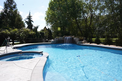 Yudaki E - Chalfont, PA - This professional installation features a multi-tier natural stone waterfall cascading into a gunite pool with a smooth, freeform design. The waterfall is constructed with large, irregularly shaped boulders creating a natural, cascading spillway that flows into the pool. The pool itself has a clean, gunite finish with a gently curving edge and integrated seating areas, surrounded by lush greenery and additional rock accents that enhance the natural aesthetic of the outdoor living space.
