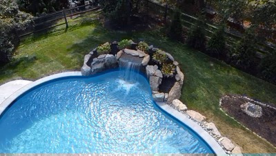 Yudaki F - Chalfont, PA - This professionally installed outdoor feature showcases a natural stone waterfall cascading gently into a gunite pool. The waterfall is constructed with large, irregularly shaped boulders that create a rustic, organic look, while the pool itself features a smooth, curved concrete edge that complements the natural materials. The surrounding landscape includes well-maintained grass, additional boulders for accenting, and a wooden fence that provides privacy and frames the serene backyard setting.