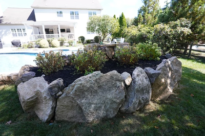 Yudaki H - Chalfont, PA - This professional installation features a multi-tiered natural stone waterfall cascading into a gunite pool. The waterfall is constructed with large, irregularly shaped boulders arranged to create a natural, flowing cascade. The pool area is bordered by additional boulders and lush landscaping, enhancing the natural aesthetic. The setting is a well-maintained backyard with a large house and mature trees in the background, providing a serene and private outdoor living space.