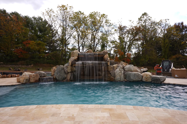 Natural stone boulder waterfall cascading into a swimming pool framed by autumn trees