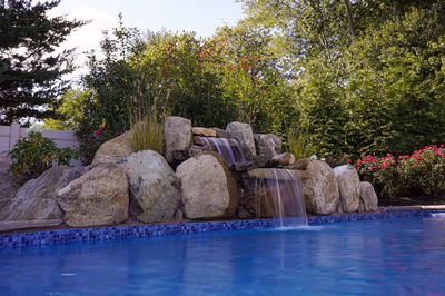2 Tier Display A - Williamstown, NJ - This professionally installed two-tier natural stone waterfall features smooth, polished boulders arranged in a cascading design, with water flowing gracefully over each tier into a gunite pool below. The waterfall's tiered structure showcases exceptional craftsmanship, blending natural stone textures with clean lines for a harmonious aesthetic. Surrounded by lush landscaping with flowering shrubs and trees, the installation creates a serene backyard oasis that enhances the outdoor living space in Williamstown, NJ.
