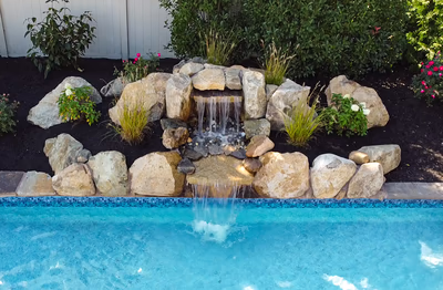 2 Tier Display B - Williamstown, NJ - This professionally installed 2-tier natural stone waterfall features a multi-level cascade that flows over carefully placed boulders into a gunite pool below. The waterfall is constructed with durable natural stone and integrated into a landscaped backyard setting, surrounded by lush greenery and decorative plants. The clean lines and quality craftsmanship create a serene water feature that enhances the outdoor living space in Williamstown, NJ.