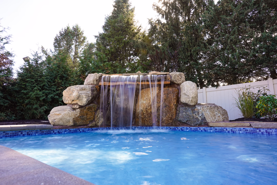 Corner Grotto A - Williamstown, NJ - This professional installation features a multi-tier natural stone waterfall cascading gracefully into a gunite pool. The waterfall is constructed with large, irregularly shaped boulders that create a rustic, organic look, while the smooth, flowing water adds a soothing element to the space. The pool has a clean, inviting blue surface with a decorative mosaic tile edge, and is surrounded by lush greenery and a white privacy fence, creating a serene backyard retreat in Williamstown, NJ.
