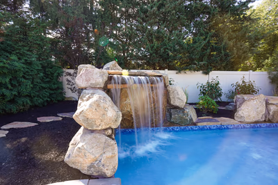 Corner Grotto B - Williamstown, NJ - This custom installation features a multi-tier natural stone waterfall cascading into a gunite pool. The waterfall is constructed from large, irregularly shaped boulders that create a rustic, organic look, with water flowing smoothly over each tier. The pool itself has a clean, blue finish and is surrounded by carefully placed landscaping, including dark mulch and additional boulders for a natural, serene setting.